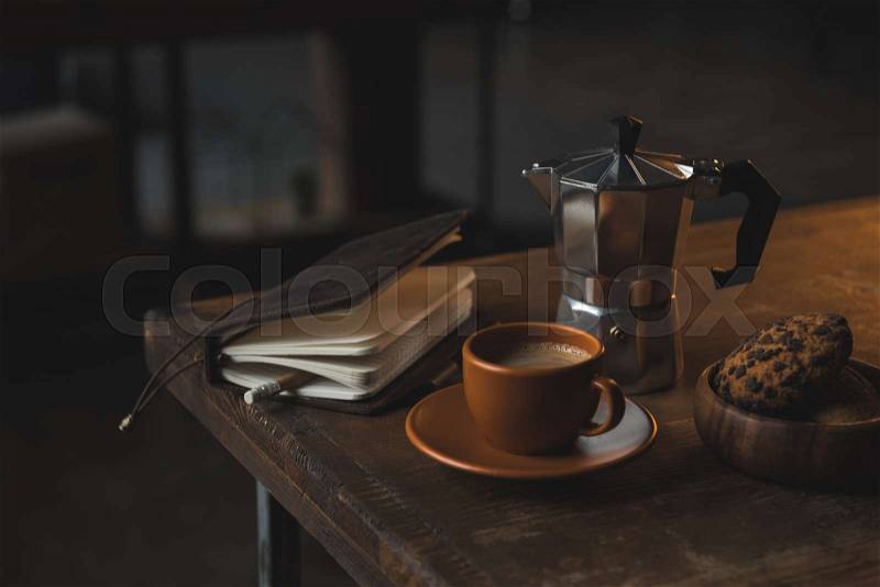 Close-up view of notebook with pencil, cup of coffee, moka pot and chocolate chip cookies on wooden table, stock photo