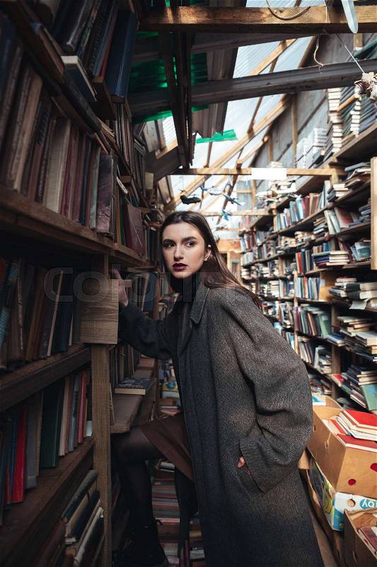 Portrait of a fashionable young woman choosing book from a shelf in a warehouse, stock photo