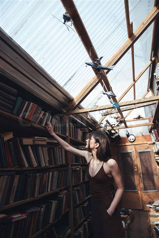 Young brunette woman wearing dress searching book in library, stock photo
