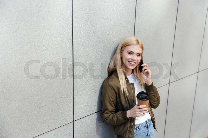 Happy young blonde woman with coffee talking on smartphone while walking in the street, stock photo