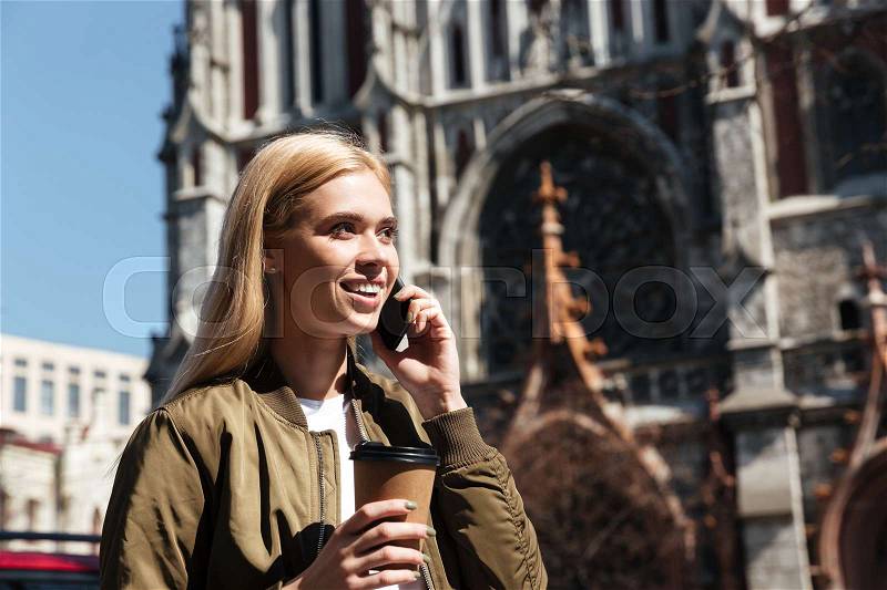 Smiling young blonde woman with coffee talking on smartphone while walking in the street, stock photo