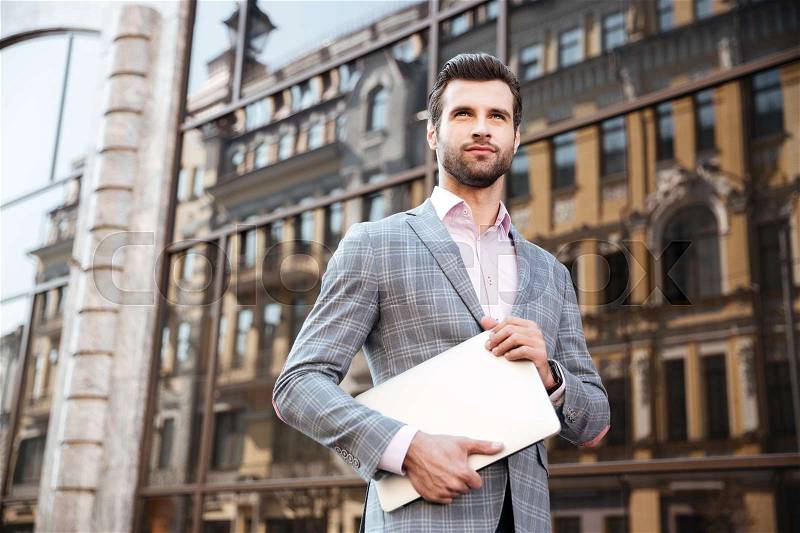 Portrait of a young serious man in jacket holding laptop computer while standing in a city area, stock photo