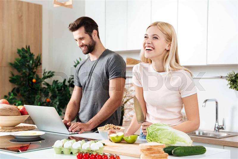 Smiling couple using notebook to cook in their kitchen and laughing, stock photo