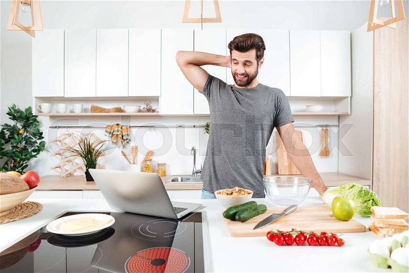 Confused handsome man cooking vegetable salad in kitchen with laptop, stock photo