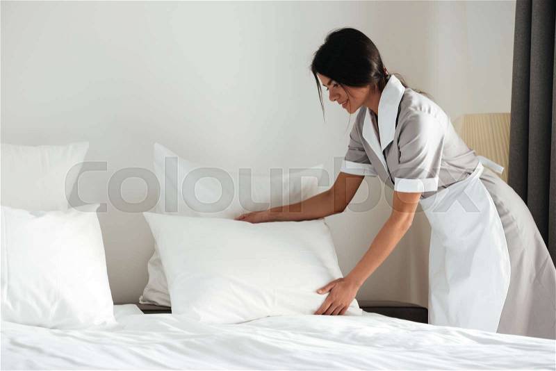 Young hotel maid setting up white pillow on bed sheet in hotel room, stock photo
