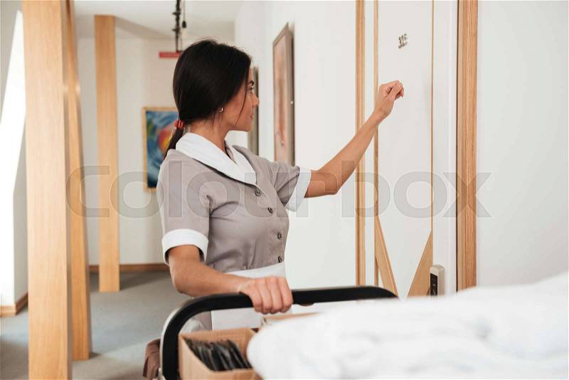 Hotel maid knocking on the hotel room door for room service holding housekeeping cart, stock photo