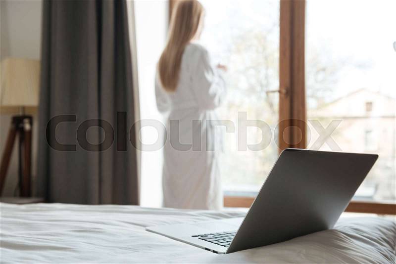 Open laptop lying on a bed with a young woman looking at the window on a background, stock photo