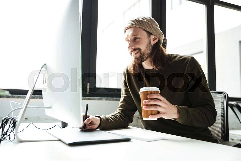 Image of young handsome man in office using computer. Looking aside drinking coffee, stock photo