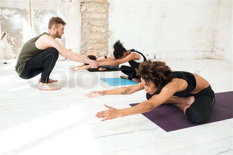 Male yoga instructor helping a woman to do yoga stretches in a studio, stock photo