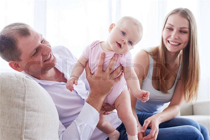 Mid shot of good-looking father holding fantastic baby while sitting with wife on sofa, stock photo
