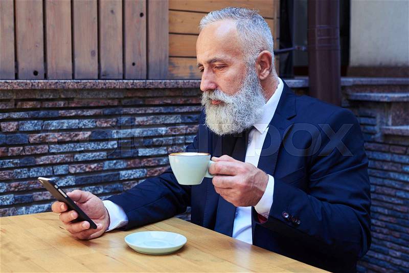 Mid shot of handsome old man drinking coffee while surfing in mobile phone, stock photo