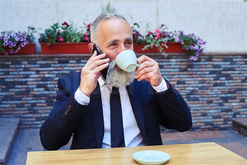 Mid shot of smiling gentleman talking on phone while drinking coffee, stock photo
