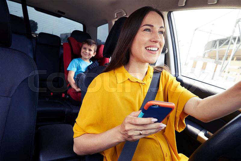 Close-up shot of a mother driving a car, buckled the seat belt, holding phone. Laughing boy on back seat, stock photo
