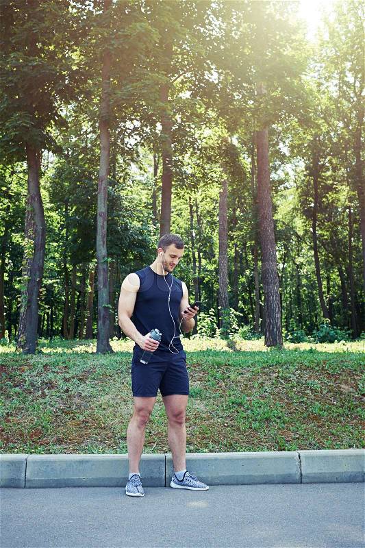 A full length image of a standing athlete looking at phone, wearing holding a bottle of water. Watching something funny, stock photo