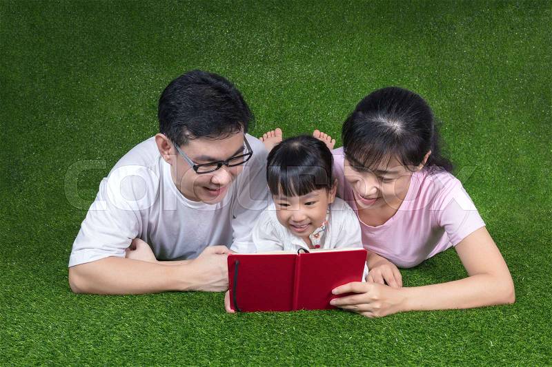 Asian Chinese parents and daughter lying on the grass and reading the book at outdoor park, stock photo