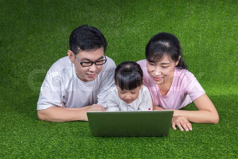 Asian Chinese parents and daughter lying on the grass and playing laptop at outdoor park, stock photo