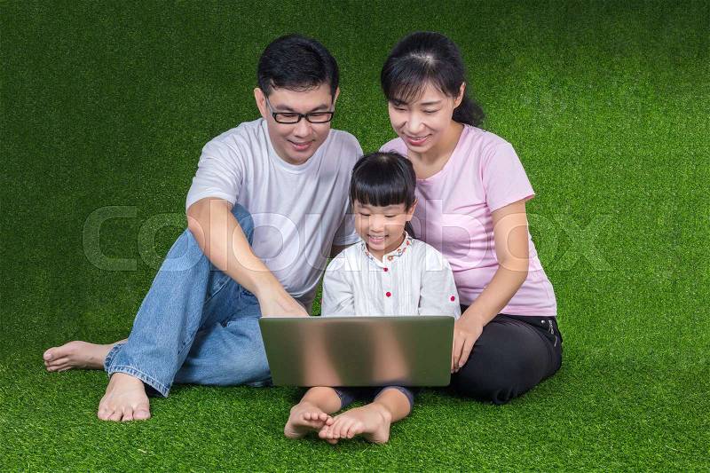 Asian Chinese parents and daughter sitting on the grass and playing laptop at outdoor park, stock photo