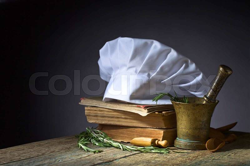 Old copper mortar with rosemary. Culinary books , chef hat and wooden spoons . Kitchen accessories on the old wooden table , stock photo