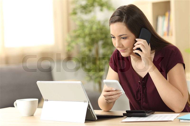 Entrepreneur working on line with phones and tablet pc sitting in a table in the living room at home with a window in the background, stock photo