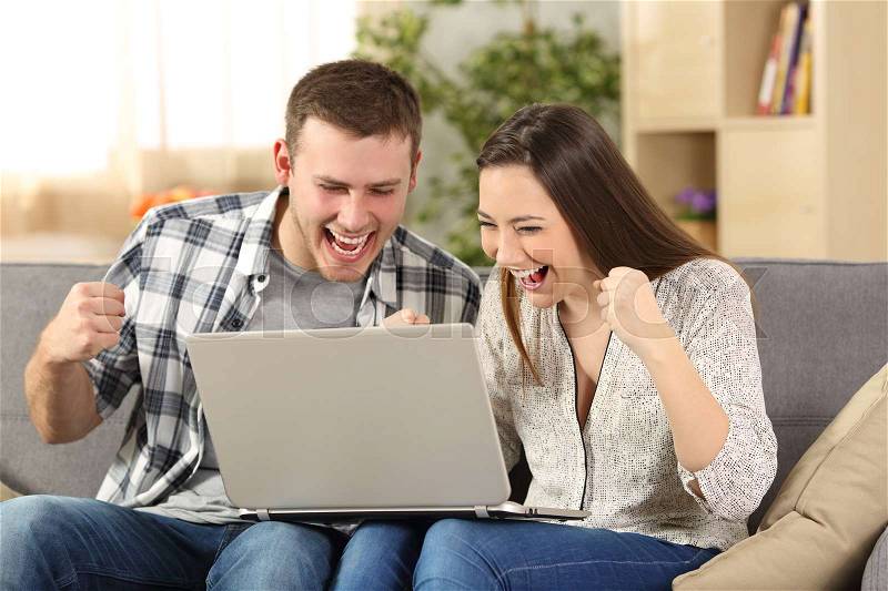 Excited couple reading news on line sitting on a couch in the living room at home, stock photo