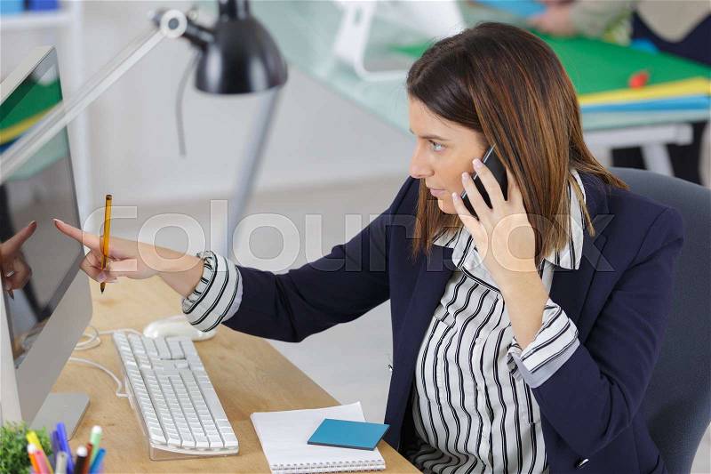 Businesswoman using landline phone while pointing towards computer in office, stock photo