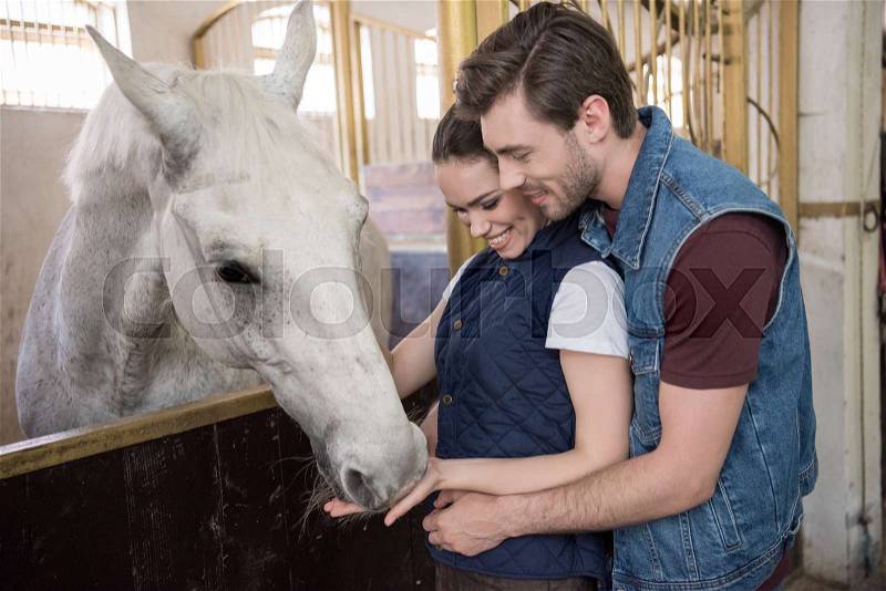 Happy young couple feeding beautiful white purebred horse, stock photo