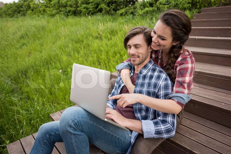 Young smiling couple using laptop while sitting on stairs at park, stock photo