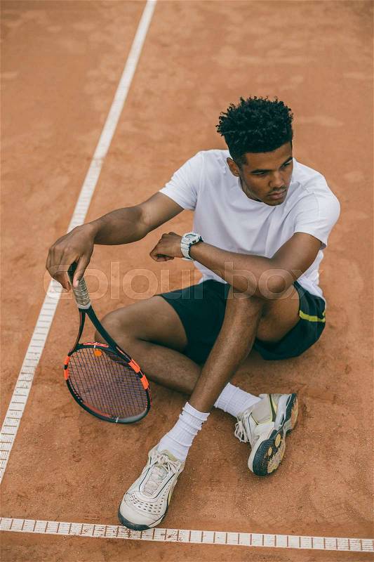 Top view young man tennis player sits on the ground of a tennis court. Serious african man relaxing after good game, stock photo
