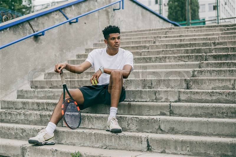 Confident young man in sports clothes on concrete steps. Man holding tennis racket and looking away while sits against grey background, stock photo
