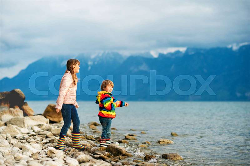 Two happy kids, little brother and big sister, playing together by lake Geneva on a very cloudy day with swiss mountains Alps on background. Image taken in Lausanne, Switzerland, stock photo