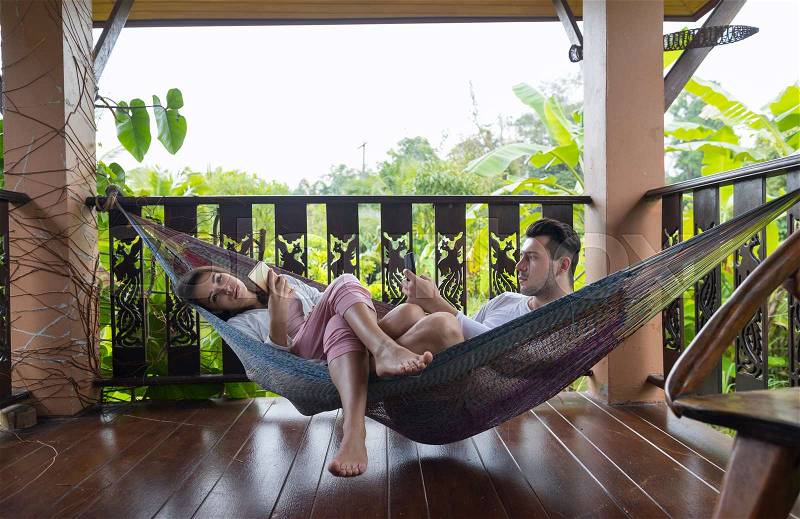 Young Couple Lying In Hammock On Terrace Tropical Hotel, Man And Woman Using Cell Smart Phone Chatting Tropic Holiday Vacation Green Forest, stock photo
