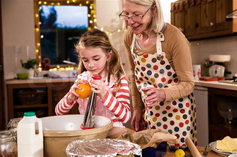 Little girl is making a cake with her grandmother. She is grating orange peel in to a bowl and her grandmother is guiding her, stock photo