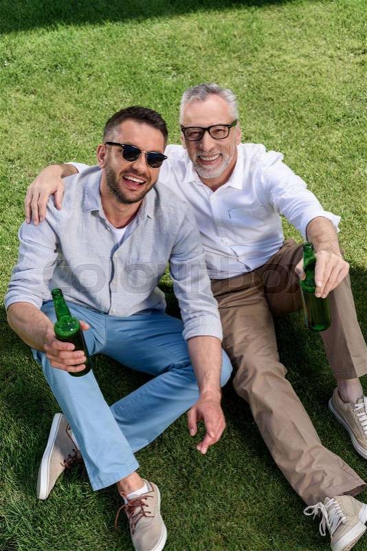 Happy father and adult son drinkig beer and sitting on green grass, stock photo