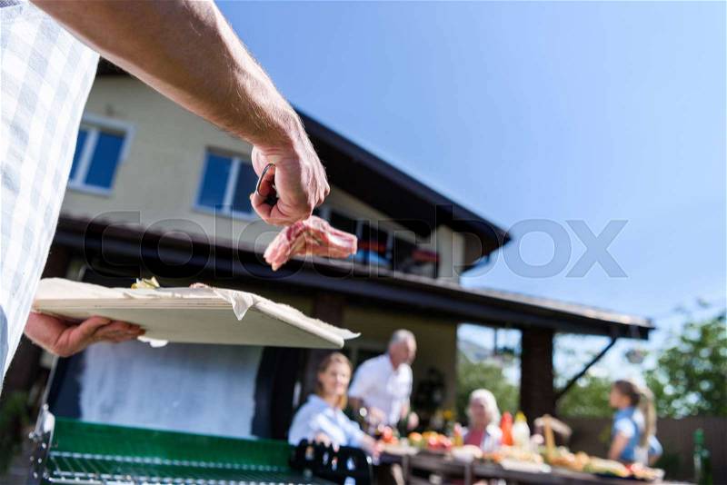 Cropped shot of man preparing meat at outdoor grill and family sitting at table outdoors, stock photo