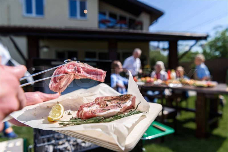 Cropped shot of man preparing meat at outdoor grill and family sitting at table outdoors, stock photo