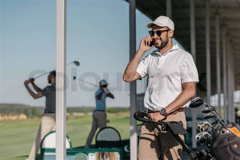 Smiling golf player talking on smartphone while friends playing golf, stock photo