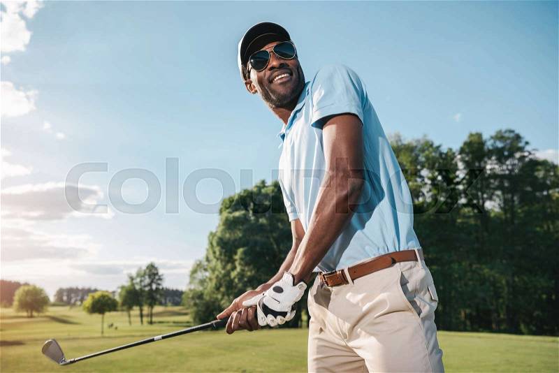 Smiling african american man in cap and sunglasses holding club and playing golf , stock photo