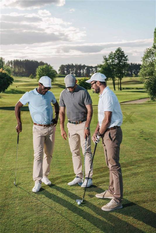 Multiethnic group of golfers holding clubs and talking while standing on green grass, stock photo