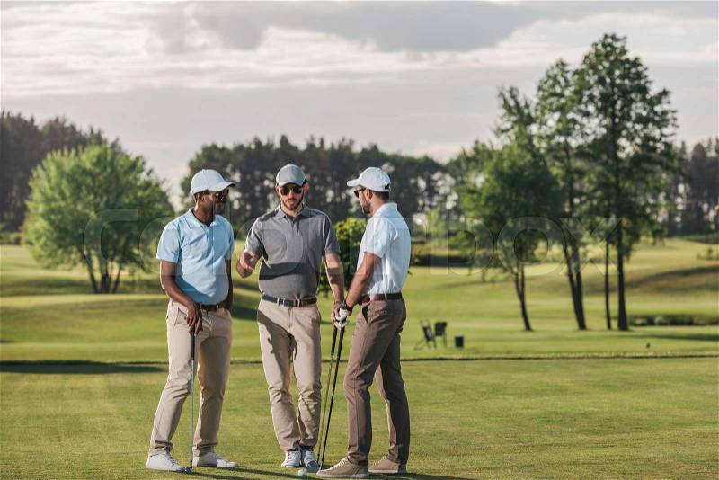 Multiethnic group of golfers holding clubs and talking while standing on green grass, stock photo