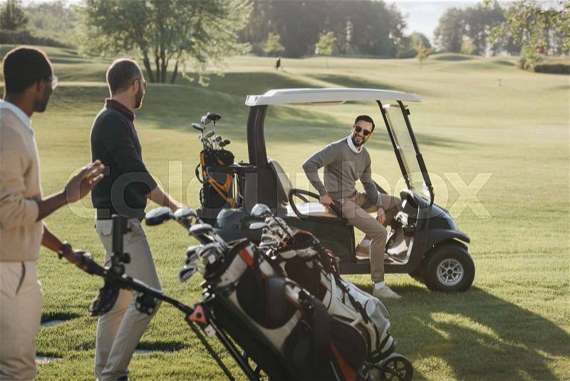 Multiethnic friends with golf bags and golf cart spending time together on golf course, stock photo