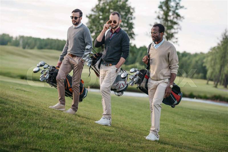 Stylish multiethnic friends holding bags with golf clubs and walking on golf course , stock photo