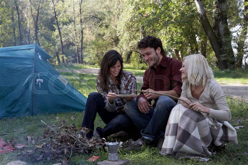 Three people at campsite cooking hot dog, stock photo
