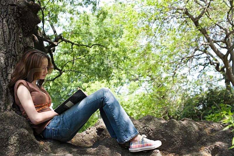 Girl writing in a notebook near a tree, stock photo