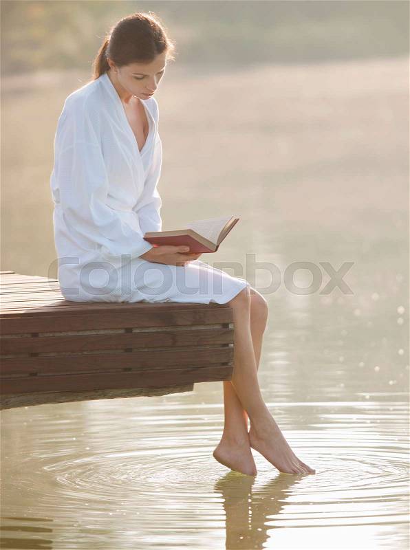 Woman reading and dangling feet in lake, stock photo