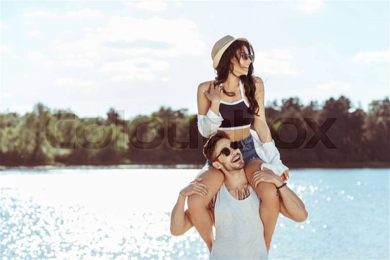 Attractive woman sitting on the shoulders of smiling man at riverside, stock photo
