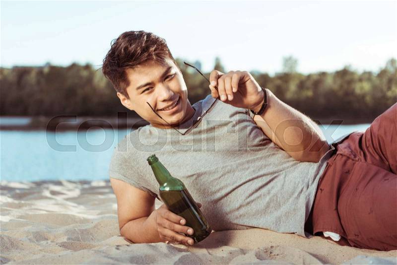 Young smiling asian man lying on sandy beach with bottle of beer and looking at camera, stock photo