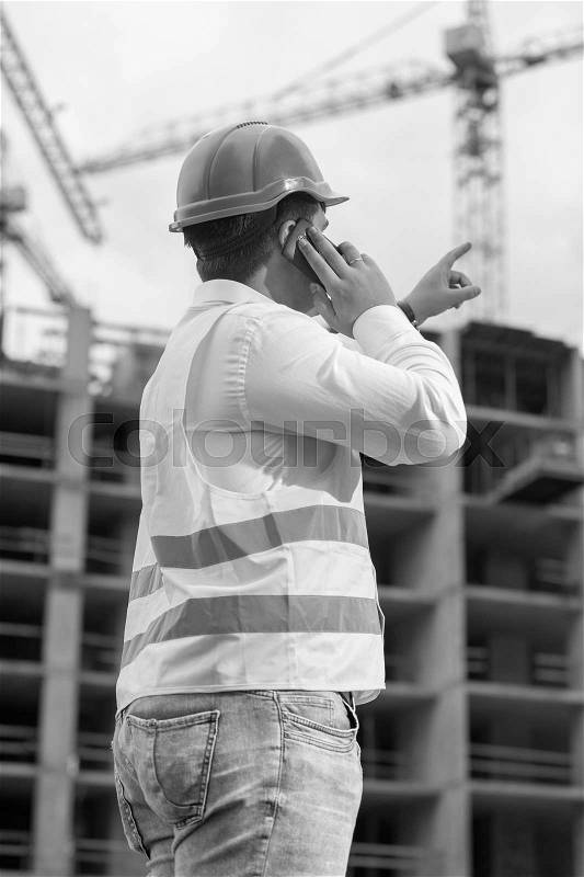 Black and white rear view image of construction engineer talking by phone and pointing at building site, stock photo