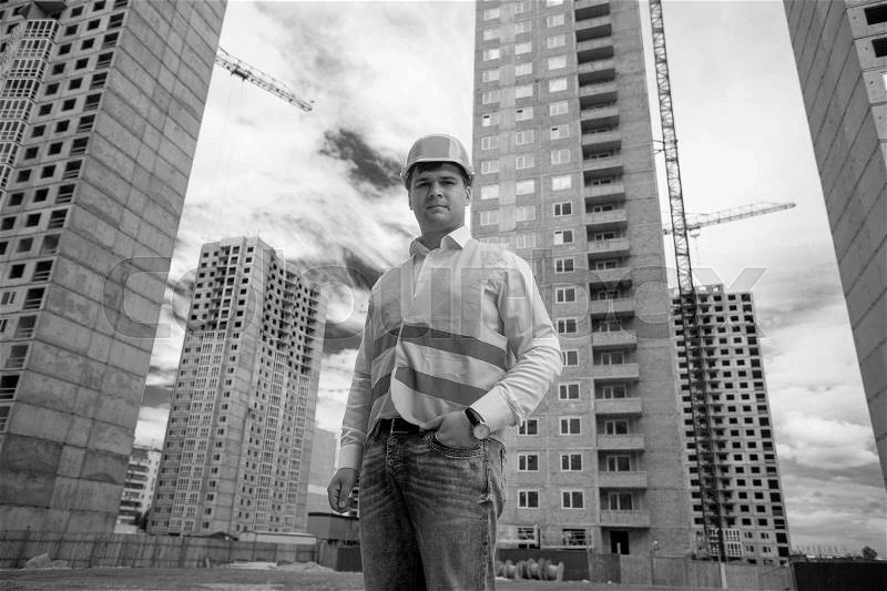 Black and white portrait of smiling engineer posing against buildings under construction, stock photo
