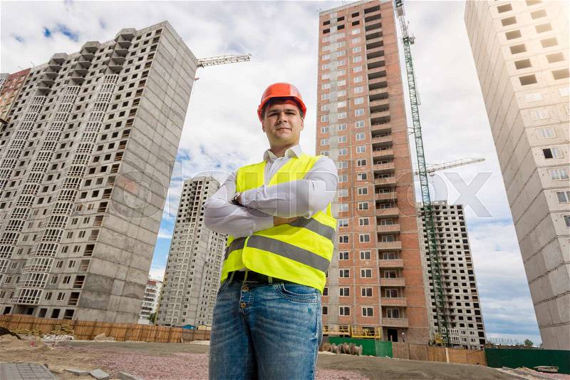 Young architect in hardhat and safety vest posing against new buildings, stock photo