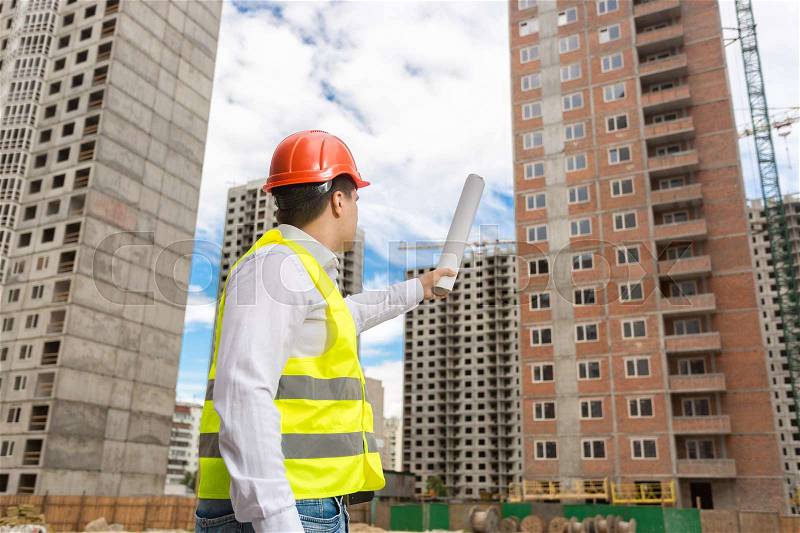 Architect in hardhat pointing with rolled blueprints at building under construction, stock photo